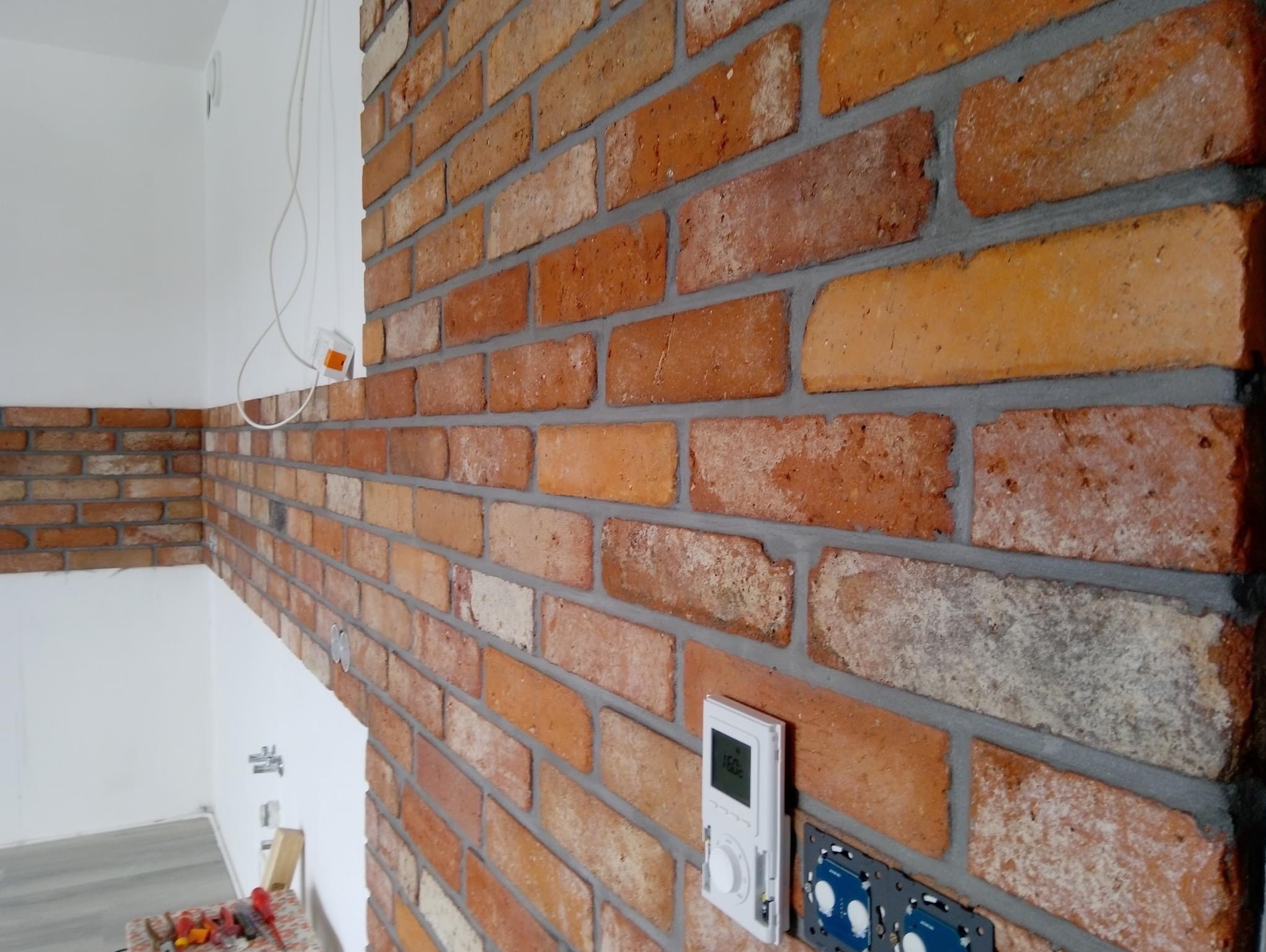 Ściana z cegły klinkierowej w odcieniach czerwieni i pomarańczy, z widocznymi fugami w kolorze szarym, zamontowany regulator temperatury i gniazdka elektryczne, fragment białej ściany w tle.