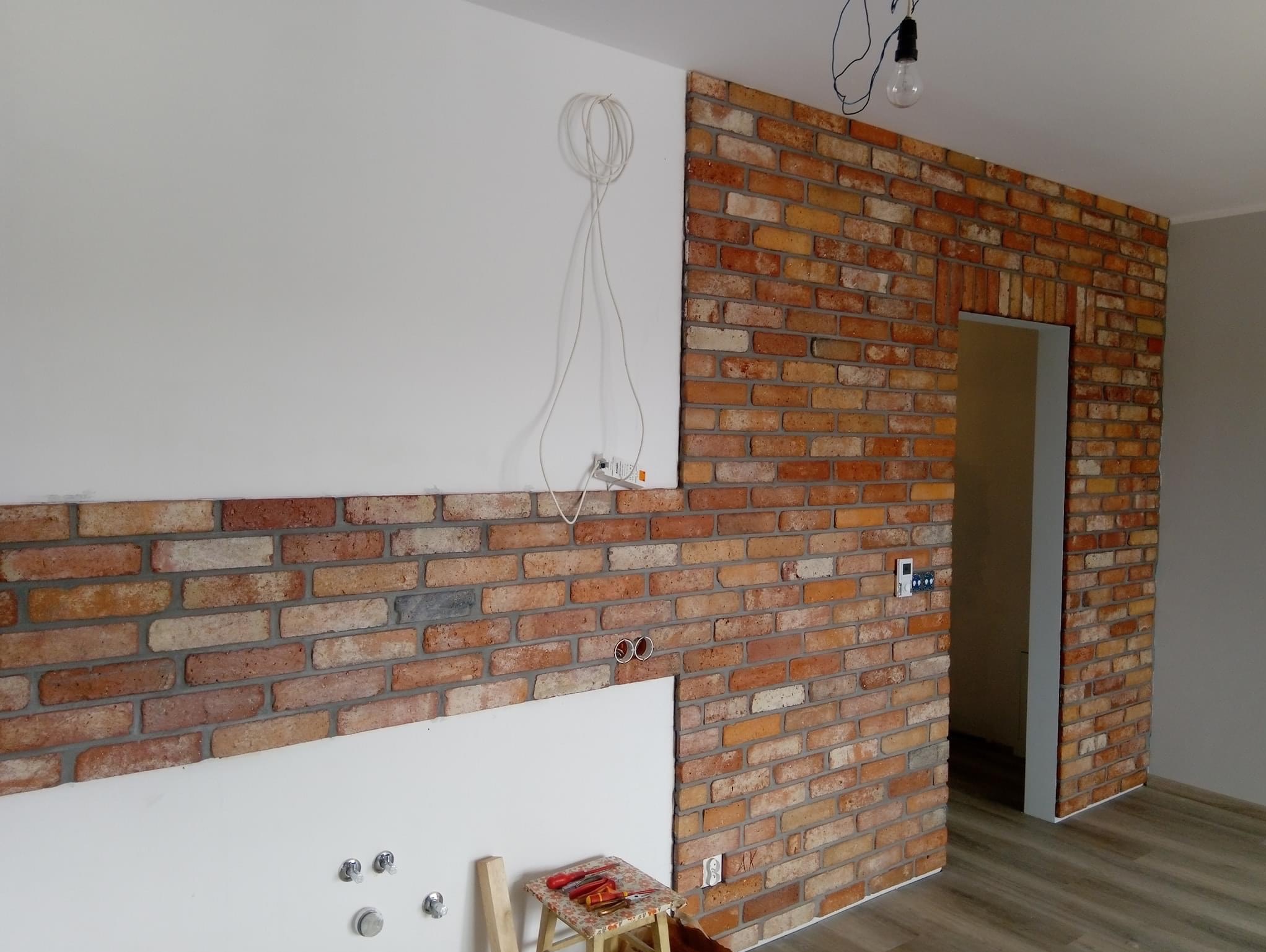 Częściowo wykończona ściana z cegły klinkierowej w odcieniach czerwieni i żółci, z widocznym otworem drzwiowym, obok fragmentu ściany przygotowanej do malowania z wystającymi kablami i gniazdkami...