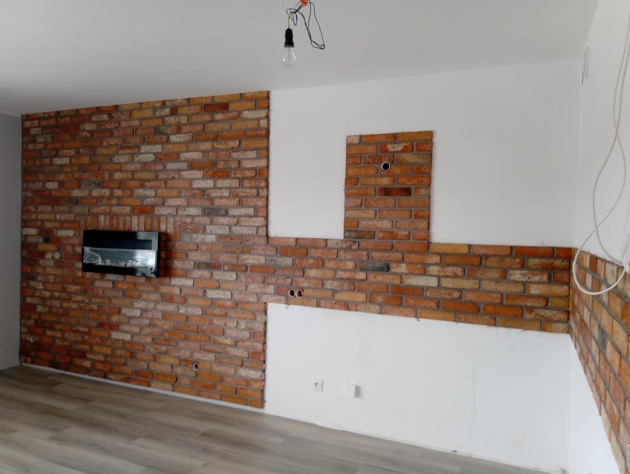 Częściowo wykończona ściana z cegły klinkierowej w salonie, widoczne przygotowane otwory pod instalacje elektryczne i fragmenty białej ściany, surowa żarówka zwisająca z sufitu.