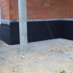 Zabezpieczone hydroizolacją fundamenty domu murowanego z cegły, widoczny fragment ściany i betonowy słup konstrukcyjny, w tle zaparkowany samochód dostawczy.