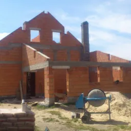 Budowa domu murowanego w trakcie: widoczne ściany z cegły, otwory okienne, komin, betonowe nadproża, betoniarka i palety z zaprawą murarską obok czerwonego samochodu dostawczego.