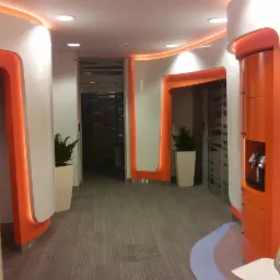 Nowoczesny korytarz biurowy z białymi ścianami i pomarańczowymi akcentami, roślinami w białych donicach i szarą wykładziną dywanową.