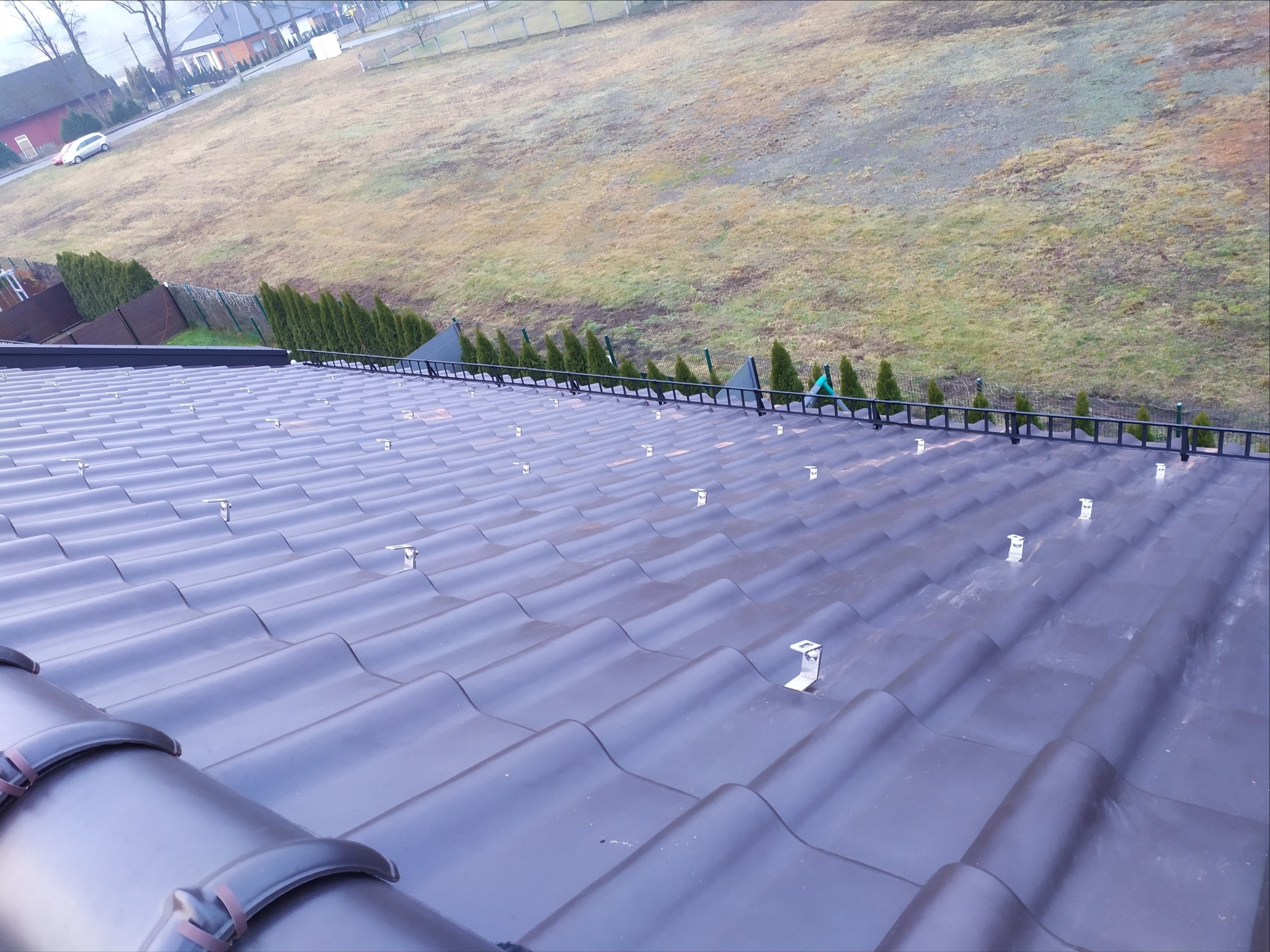 Widok z góry na ciemną, ceramiczną dachówkę z zamontowanymi uchwytami montażowymi pod panele słoneczne, z widocznym fragmentem instalacji odgromowej i rozległym terenem zielonym w tle.