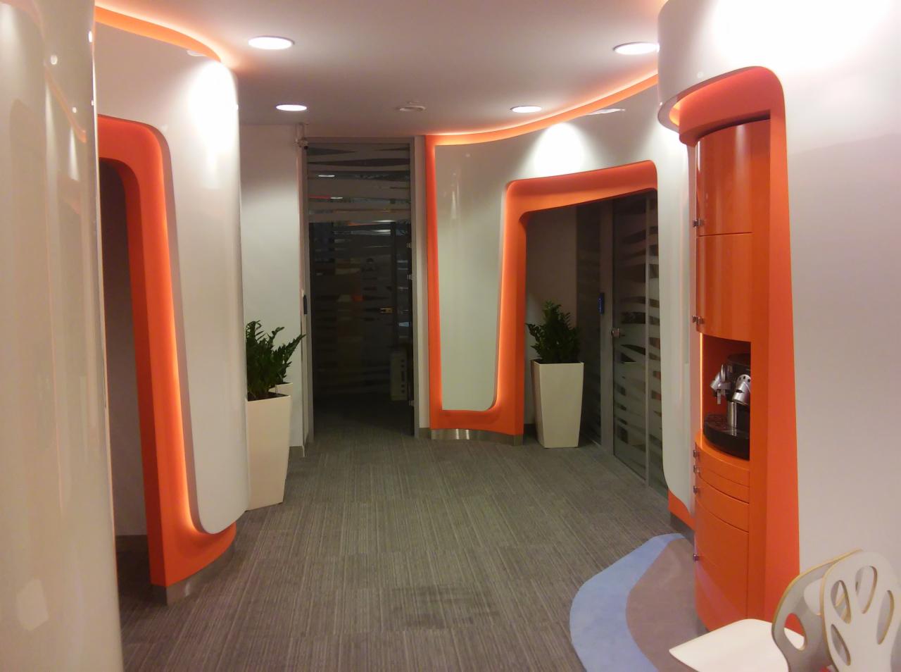 Nowoczesny korytarz biurowy z białymi ścianami i pomarańczowymi akcentami, roślinami w białych donicach i szarą wykładziną dywanową.