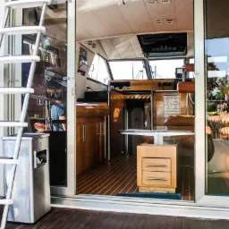 Wnętrze luksusowego jachtu motorowego z drewnianą podłogą i meblami, widoczne schody wejściowe oraz kokpit z fotelami kapitańskimi i panelem sterowania, z zewnątrz widoczny fragment zielonego węża.