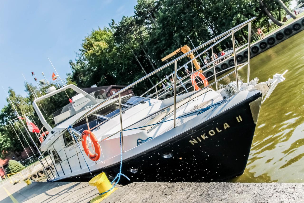 Czarny jacht motorowy 'Nikola II' zacumowany przy betonowym nabrzeżu, z pomarańczową boją i liną cumowniczą, widok na zielone drzewa i spacerujących ludzi w tle.