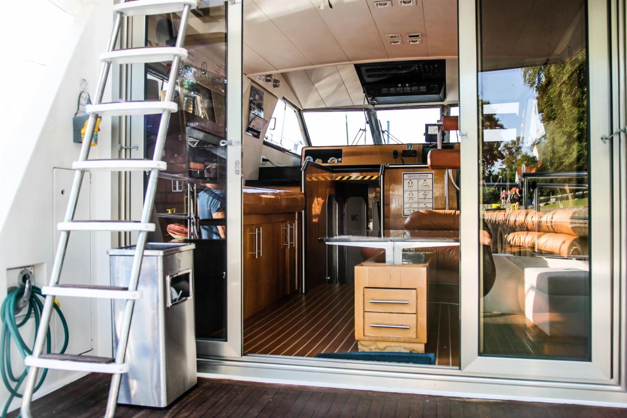 Wnętrze luksusowego jachtu motorowego z drewnianą podłogą i meblami, widoczne schody wejściowe oraz kokpit z fotelami kapitańskimi i panelem sterowania, z zewnątrz widoczny fragment zielonego węża.