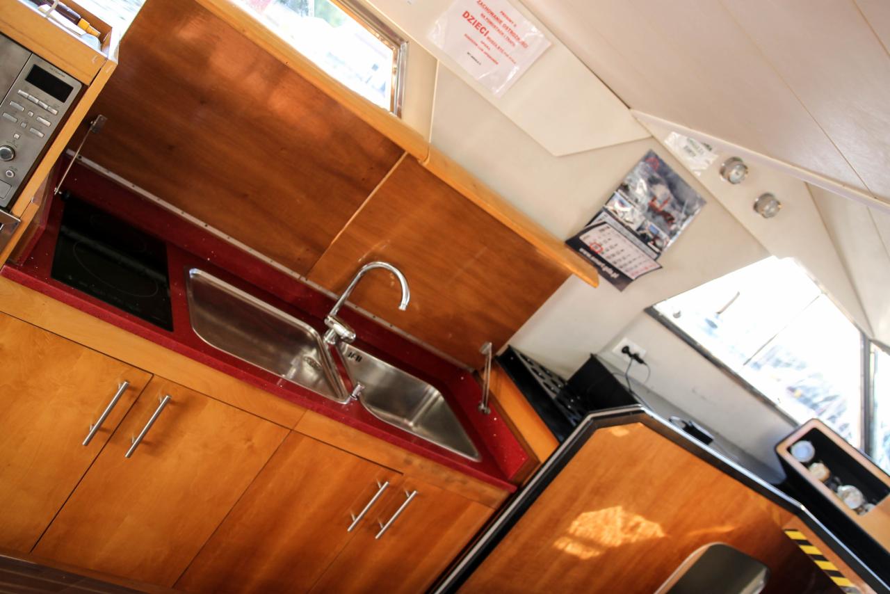 Wnętrze jachtu: zabudowa kuchenna z drewnianymi szafkami, dwukomorowym zlewem i płytą indukcyjną, widoczny kalendarz na ścianie.