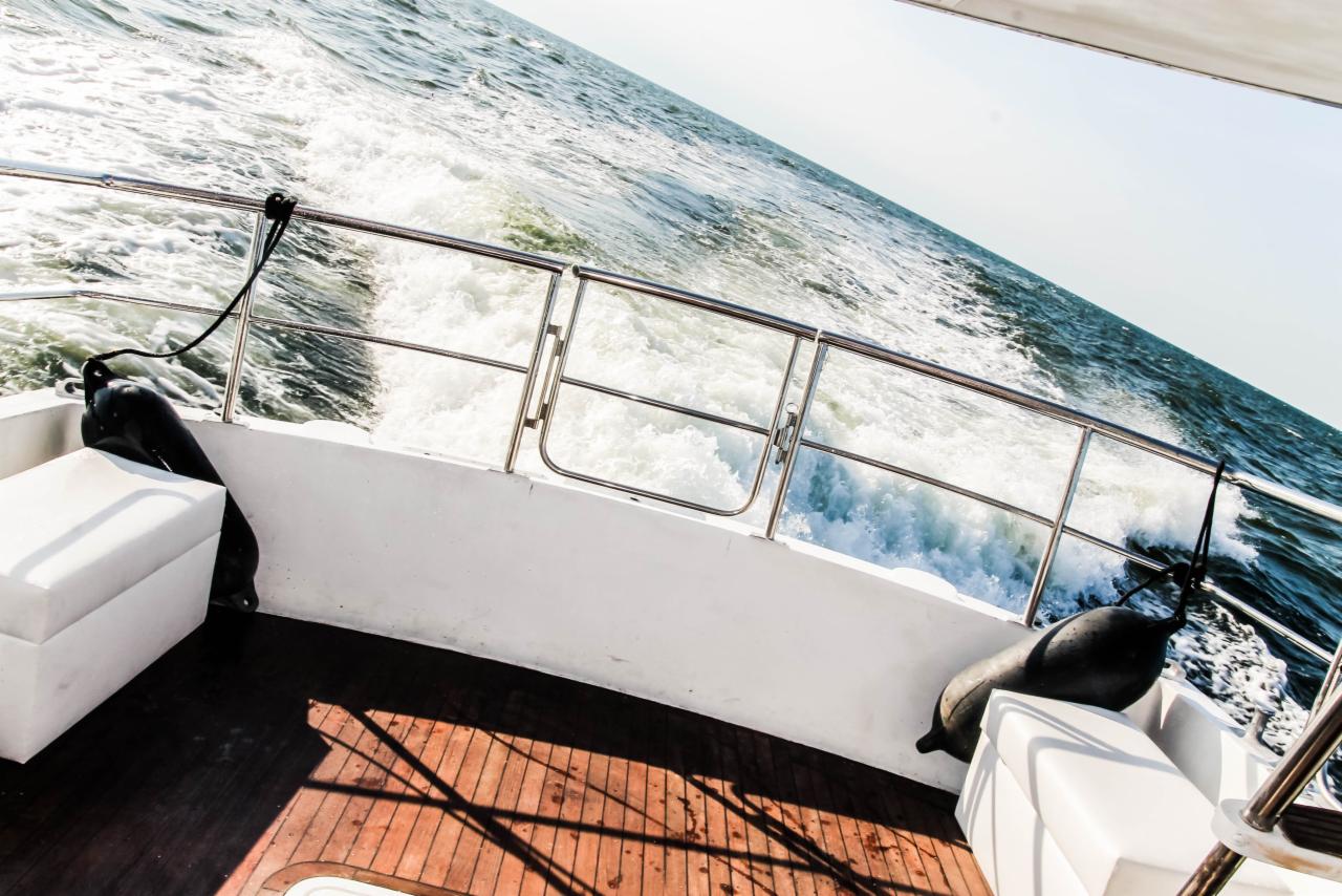 Widok z rufy jachtu motorowego na wzburzone morze, biała piana za burtą, drewniany pokład z odbiciami promieni słonecznych, czarne odbijacze przymocowane do relingu.