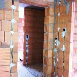 Elektryk Dominik - Montaż przewodów elektrycznych w bruzdach ceglanych ścian, zabezpieczonych gipsem. Widoczne otwory pod gniazdka, kable na podłodze, surowy stan budowy.