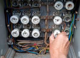 Otwarta skrzynka z bezpiecznikami i kolorowymi przewodami, dłoń w prawym dolnym rogu. Widoczne ślady korozji i zużycia na elementach.