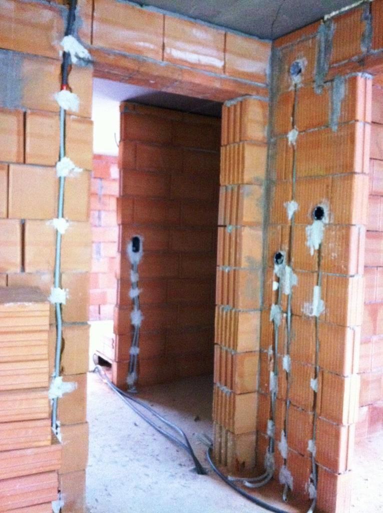 Montaż przewodów elektrycznych w bruzdach ceglanych ścian, zabezpieczonych gipsem. Widoczne otwory pod gniazdka, kable na podłodze, surowy stan budowy.