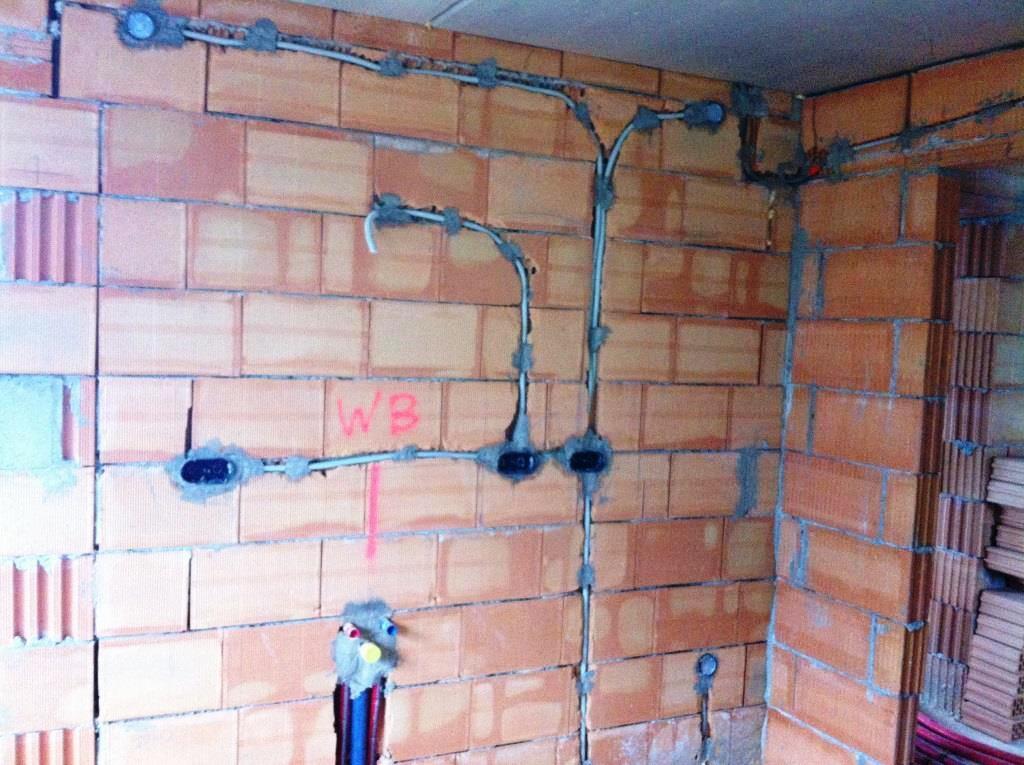 Instalacja elektryczna w surowym murze z czerwonej cegły. Widoczne przewody w peszlach, puszki elektryczne zalane gipsem, pionowa linia i napis 'WB' na ścianie.