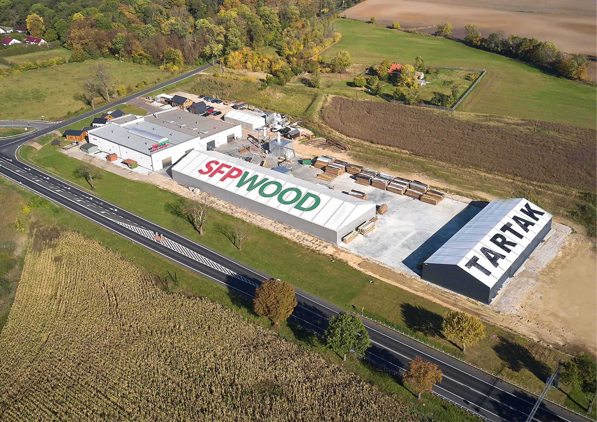 Widok z lotu ptaka na kompleks budynków firmy zajmującej się obróbką drewna, z widocznymi magazynami, budynkiem biurowym, placem składowym z ułożonym drewnem i napisem reklamowym na dachu magazynu.