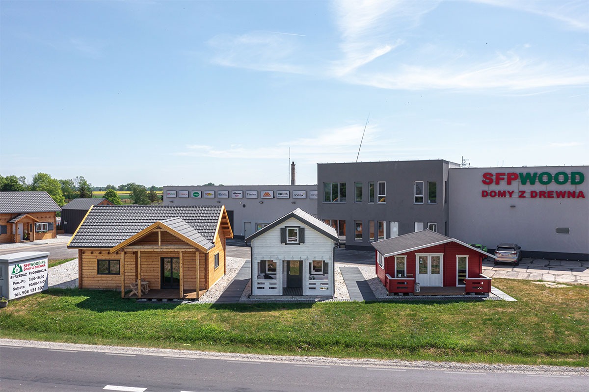 Ekspozycja trzech małych domków o różnej kolorystyce (drewniany, biały, czerwony) z ciemnymi dachami, prezentowanych przed budynkiem firmy SFPWOOD Domy z Drewna w słoneczny dzień.