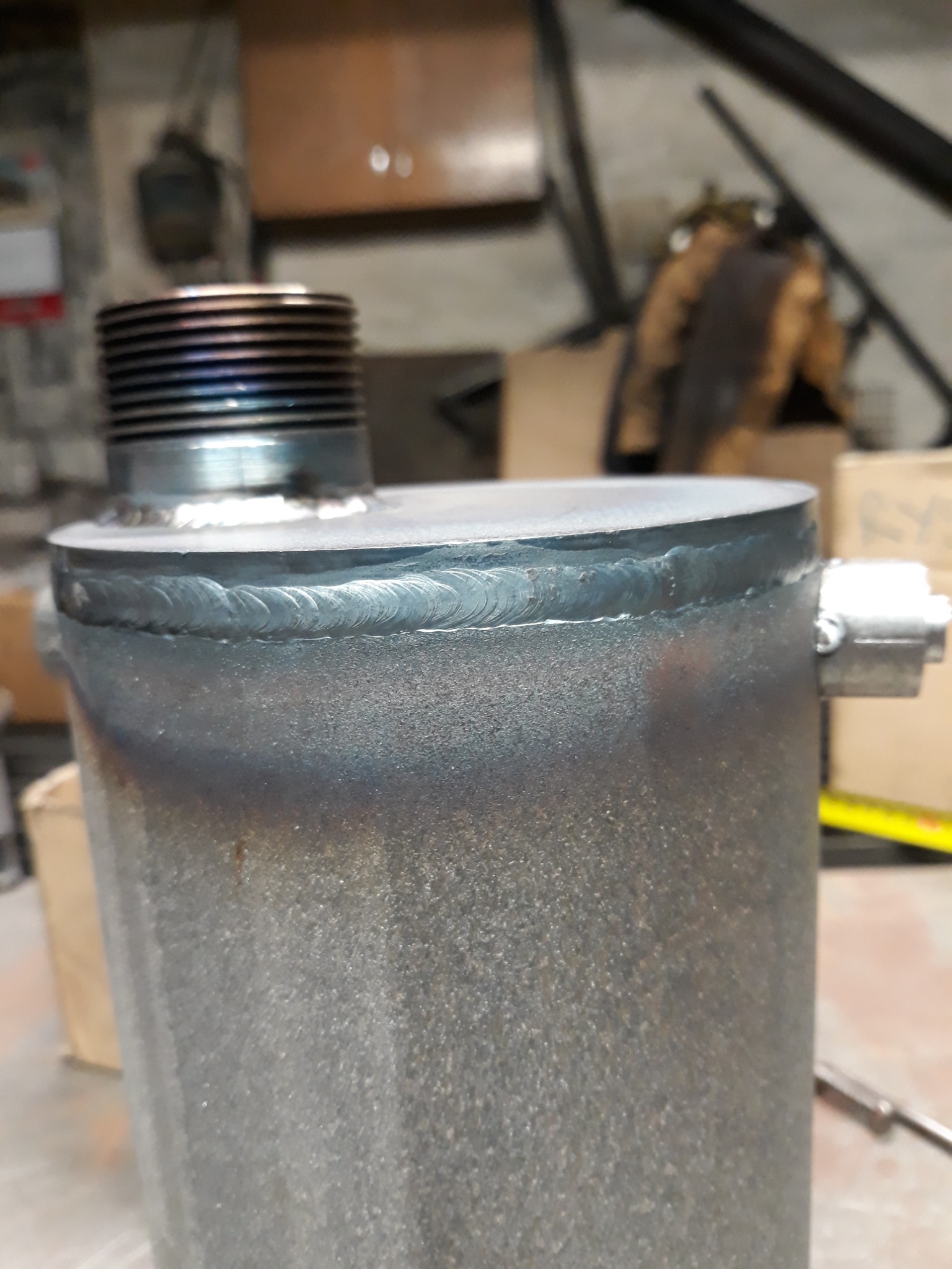 Metalowy zbiornik z widocznym spawem na górnej krawędzi, z gwintowanym króćcem na górze, ślady przypaleń po procesie obróbki termicznej.