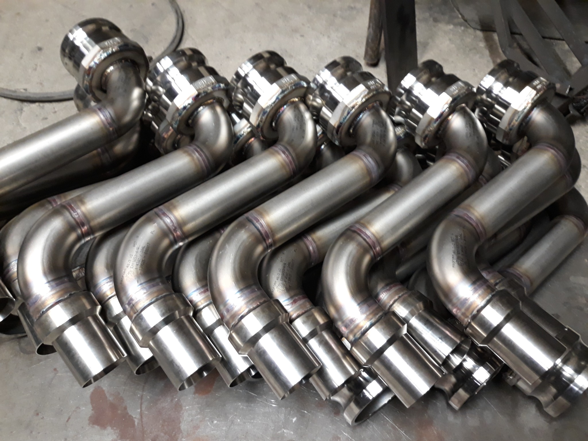 Rząd identycznych, spawanych, metalowych rur wydechowych z kołnierzami, ułożonych na szarej powierzchni, widoczne ślady spawania i oznaczenia na metalu.
