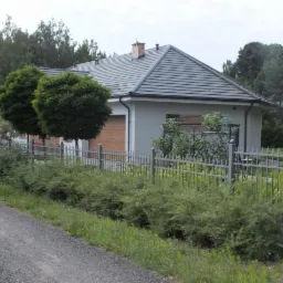 Szary dom jednorodzinny z ciemną dachówką, drewnianą bramą garażową i metalowym ogrodzeniem, otoczony zielenią. Widok z poziomu gruntu.