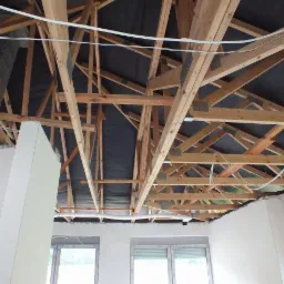 Drewniana konstrukcja dachu z membraną, widoczna od wewnątrz budynku w trakcie budowy, z fragmentami otynkowanych ścian i instalacją elektryczną.