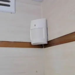Biały czujnik ruchu zamontowany w rogu pomieszczenia z kremowymi płytkami i brązowym dekoracyjnym pasem. Widoczny także fragment kratki wentylacyjnej.