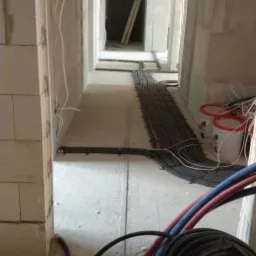 Wnętrze w trakcie instalacji ogrzewania podłogowego i instalacji elektrycznej: rury grzewcze w kolorze czerwonym i niebieskim zwinięte w kręgi na podłodze, czarne peszle elektryczne ułożone wzdłuż...