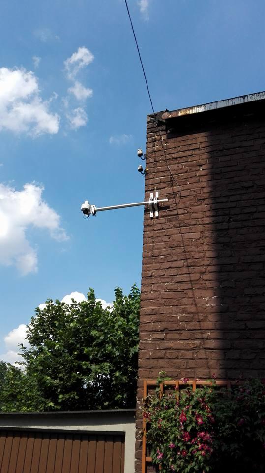 Kamera monitoringu zamontowana na brązowej, ceglanej ścianie budynku w Rudzie Śląskiej, widok z dołu na tle błękitnego nieba z chmurami.