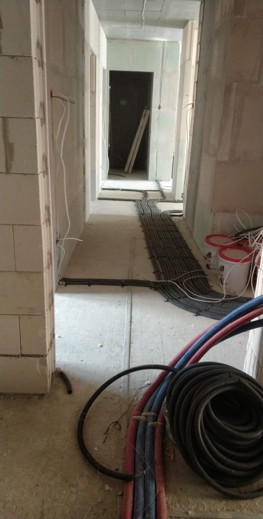 Wnętrze w trakcie instalacji ogrzewania podłogowego i instalacji elektrycznej: rury grzewcze w kolorze czerwonym i niebieskim zwinięte w kręgi na podłodze, czarne peszle elektryczne ułożone wzdłuż...