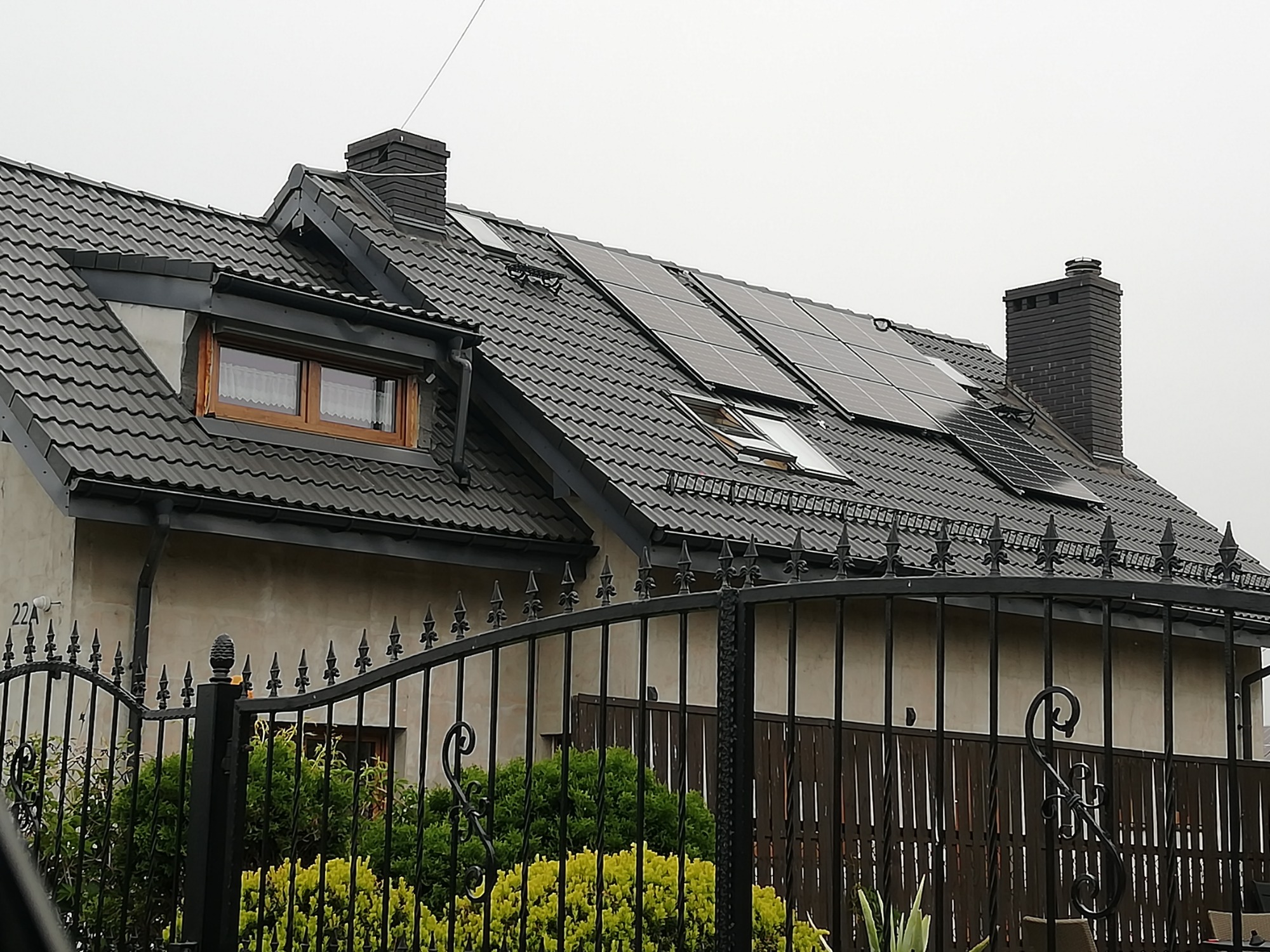 Dom jednorodzinny z szarą dachówką i panelami słonecznymi na dachu, widoczny zza czarnego, ozdobnego ogrodzenia.