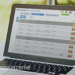 Laptop MacBook Air wyświetla panel administracyjny Clear Channel z listą powierzchni reklamowych i opcjami wyboru, na biurku. Znak 'Rozwiązanie' w lewym dolnym rogu.