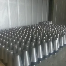 Rząd metalowych, stożkowych elementów z czarnymi podstawami, ułożonych w rzędach na metalowej powierzchni w hali produkcyjnej. Elementy po obróbce.