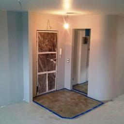 Projekt RS - Pokój w trakcie malowania na szaro, zabezpieczone podłogi i drzwi wejściowe folią malarską, widoczny fragment zaokrąglonej ściany i niedokończone prace wykończeniowe.