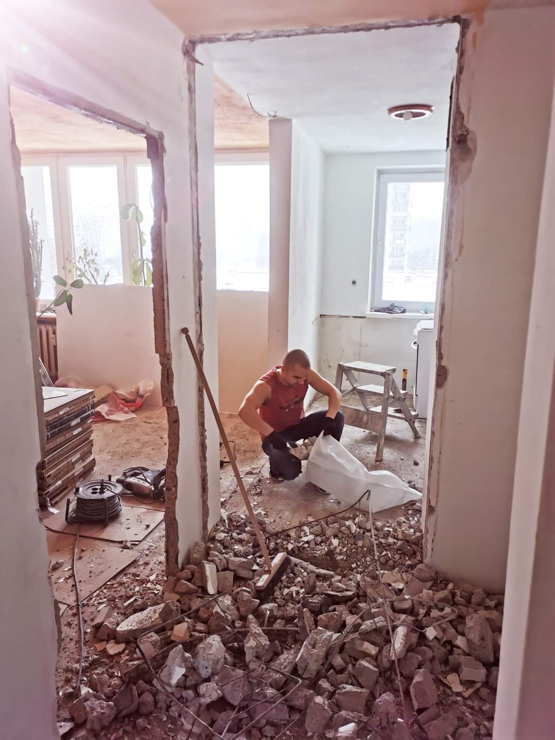 Mężczyzna w czerwonej koszulce zbiera gruz do worka podczas demontażu ściany działowej w trakcie remontu mieszkania, widoczne narzędzia i sterta płytek.