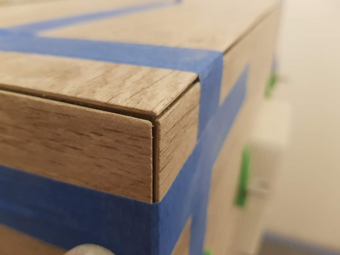 Szczegółowe ujęcie narożnika drewnianej szafki zabezpieczonej niebieską taśmą malarską, widoczne delikatne niedoskonałości połączenia elementów.