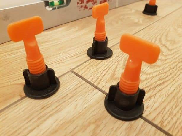 Pomarańczowe kliny poziomujące płytki podłogowe, czarne podstawki, poziomica w tle, drewnopodobne płytki w trakcie układania.