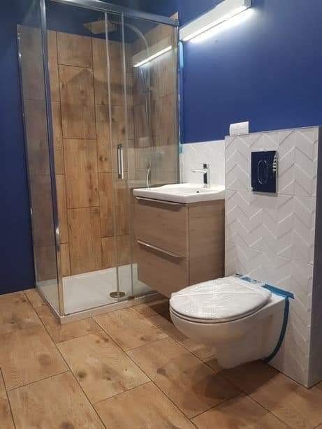 Nowoczesna łazienka z kabiną prysznicową z drewnopodobnymi płytkami, wiszącą toaletą z zabezpieczeniem folią i umywalką z szafką w odcieniach beżu na podłodze z płytek imitujących drewno.