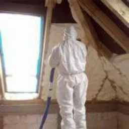 Pracownik w kombinezonie ochronnym aplikuje izolację natryskową na poddaszu, widoczne krople piany na ścianach i elementach konstrukcyjnych dachu.