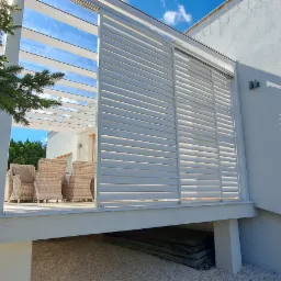 Nowoczesny taras z białą pergolą i żaluzjami przeciwsłonecznymi, rattanowe meble ogrodowe, widok z zewnątrz na jasny dzień.