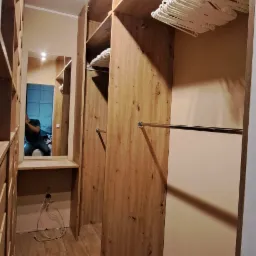 Wnętrze garderoby z drewnianymi półkami i drążkami, widoczne białe wieszaki, lustro z odbiciem osoby, drewnopodobna podłoga i zielony puf.