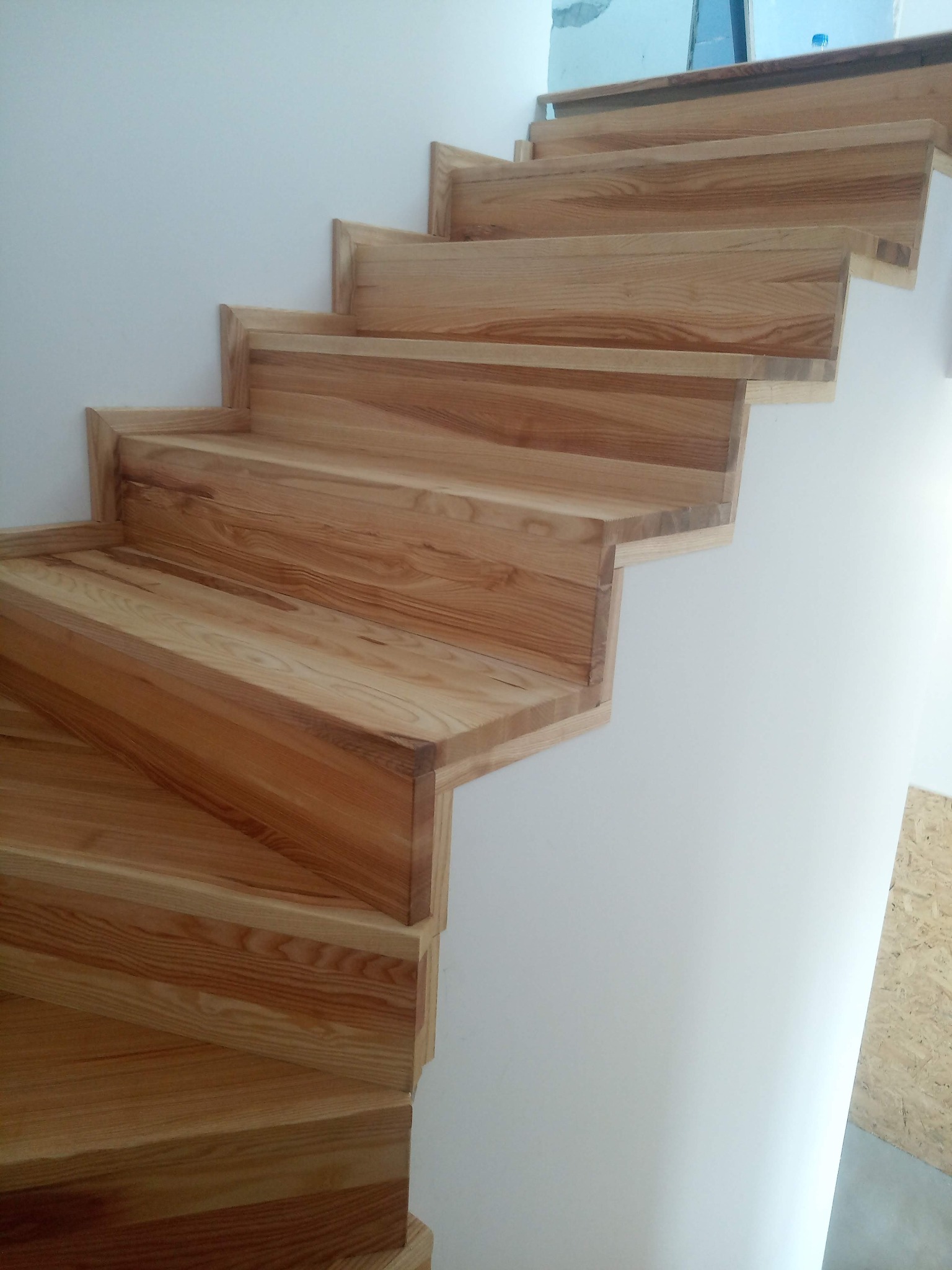 Surowe drewniane schody z widocznym usłojeniem, przylegające do białej ściany w trakcie montażu, widoczne elementy wykończeniowe pod schodami.