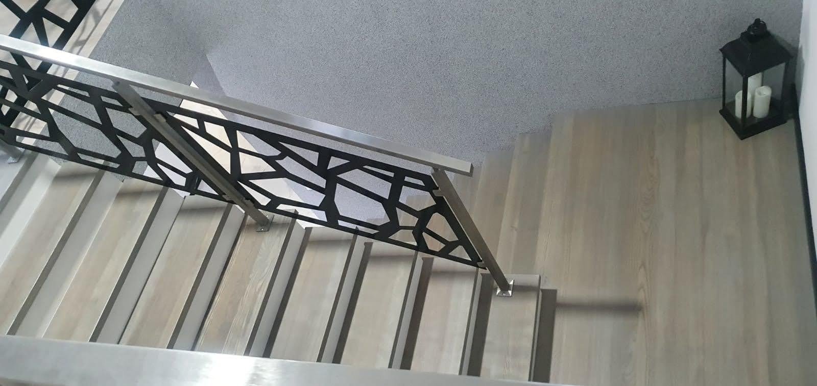 Widok z góry na jasne schody z drewnianymi stopniami i nowoczesną, metalową balustradą o geometrycznym wzorze, obok czarny lampion ze świecami.