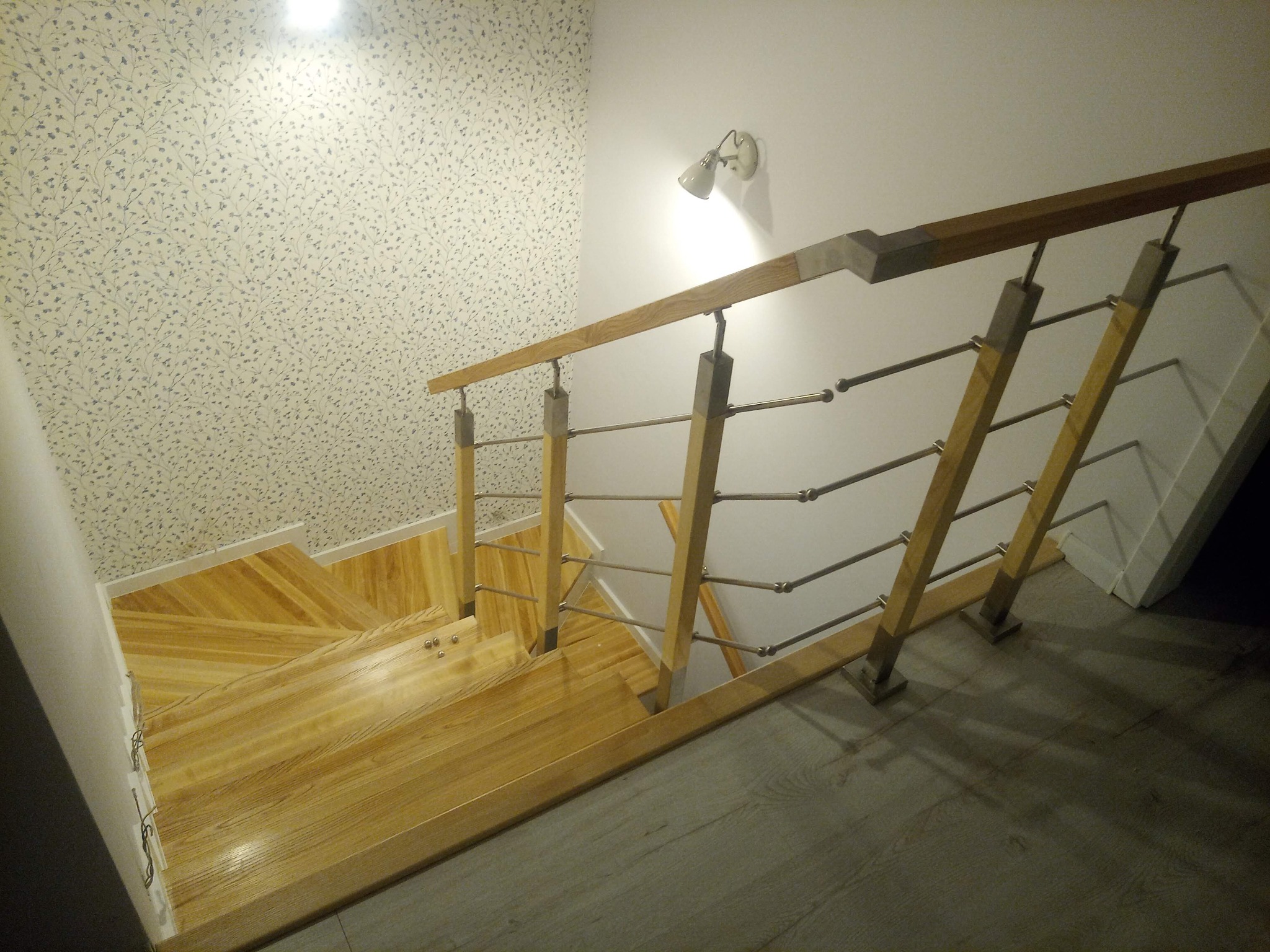 Jasne drewniane schody z metalowymi relingami, widok z góry, ściana z tapetą w drobne kwiatki, oświetlenie punktowe.