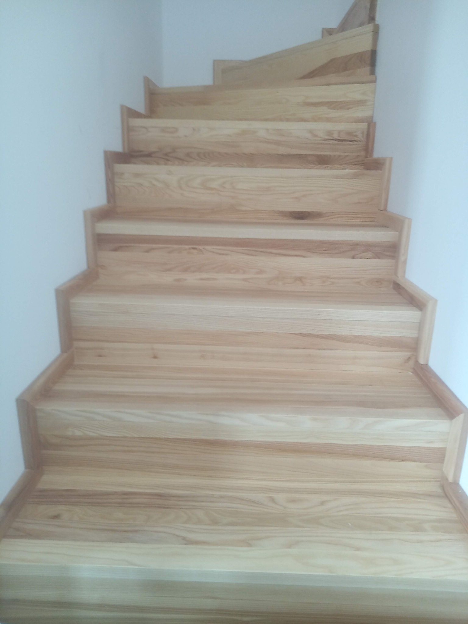 Jasne, drewniane schody z widocznym rysunkiem słojów, prowadzące w górę, przy białej ścianie.