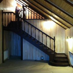 Drewkomplet Spółka z Ograniczoną Odpowiedzialnością - Drewniane schody z balustradą w domu z boazerią, oświetlone lampą. Na schodach stoi osoba. Widoczna konstrukcja sufitu i podłoga. Kątowe ujęcie.