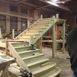 Drewkomplet Spółka z Ograniczoną Odpowiedzialnością - Surowe drewniane schody z podestem w warsztacie, w trakcie montażu. Widoczne narzędzia i elementy konstrukcyjne. Naturalne drewno, ciepłe oświetlenie.
