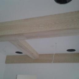 Drewkomplet Spółka z Ograniczoną Odpowiedzialnością - Dekoracyjne belki drewniane na białym suficie, z widocznymi otworami na oświetlenie. Minimalistyczny design, jasne drewno z wyraźnym usłojeniem.