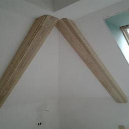 Drewkomplet Spółka z Ograniczoną Odpowiedzialnością - Dwie drewniane belki stropowe, zamontowane pod kątem na białej ścianie poddasza, z oknem dachowym w tle. Naturalny odcień drewna i widoczne słoje.