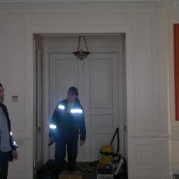 Drewkomplet Spółka z Ograniczoną Odpowiedzialnością - Montaż drzwi wewnętrznych: Dwóch monterów w odblaskowych kurtkach instaluje białe, klasyczne drzwi w jasnym pomieszczeniu z oknami i dekoracyjnymi panelami ściennymi.