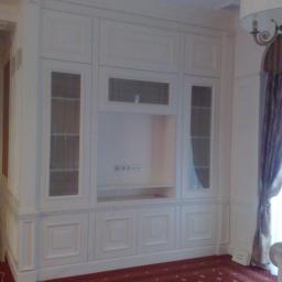 Drewkomplet Spółka z Ograniczoną Odpowiedzialnością - Biała, zabudowana szafa wnękowa z przeszklonymi witrynami i miejscem na telewizor, wkomponowana w klasyczne wnętrze z czerwonym dywanem.