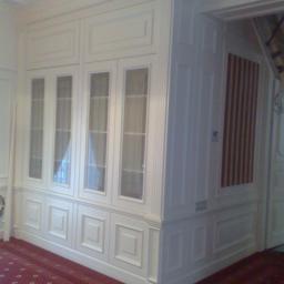 Drewkomplet Spółka z Ograniczoną Odpowiedzialnością - Biała, zabudowana szafa z przeszklonymi drzwiami, wkomponowana w ścianę z dekoracyjnymi panelami. Czerwony dywan z wzorem na podłodze, klasyczny wystrój.