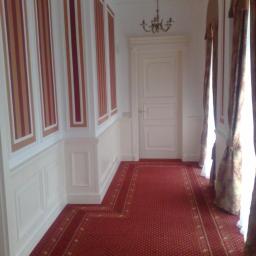 Drewkomplet Spółka z Ograniczoną Odpowiedzialnością - Korytarz z czerwonym dywanem, białymi panelami ściennymi z bordowo-złotymi pasami i klasycznymi, białymi drzwiami z ościeżnicą. Nad drzwiami wisi żyrandol.